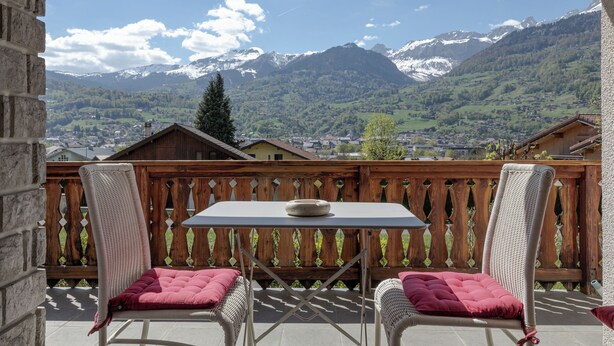 En balkon med fletstole og et bord, med udsigt over et bjergrigt landskab med huse og grønt.