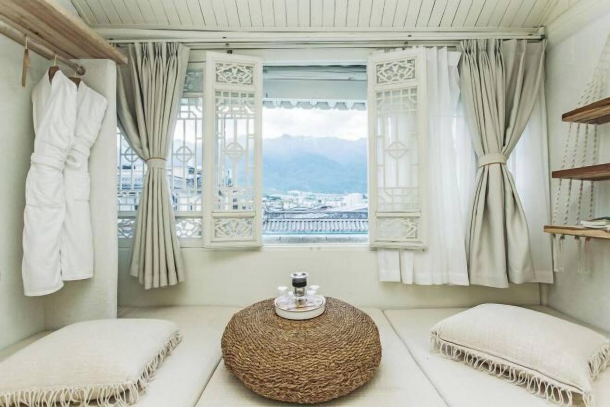 A room with a view of a city and mountains, a round woven ottoman, and two cushioned seating areas.