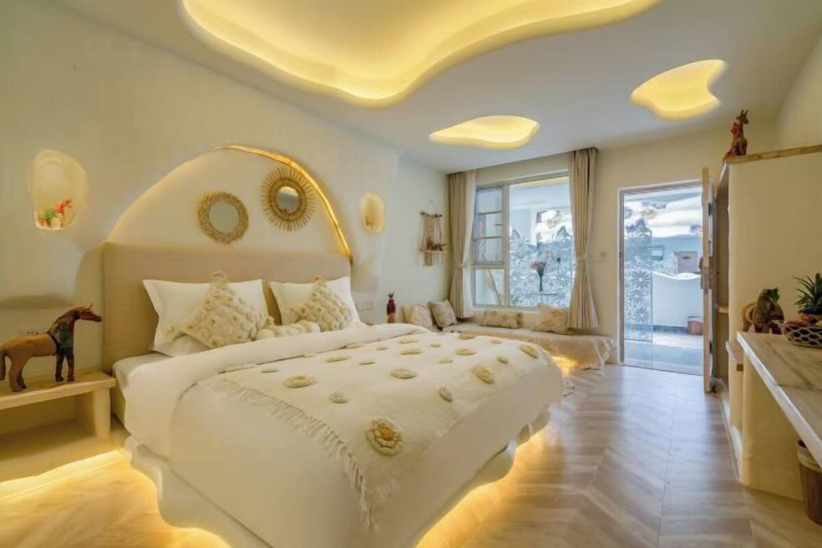 A bedroom with a large bed, a bedside table, a window with curtains, and a view of the outside.