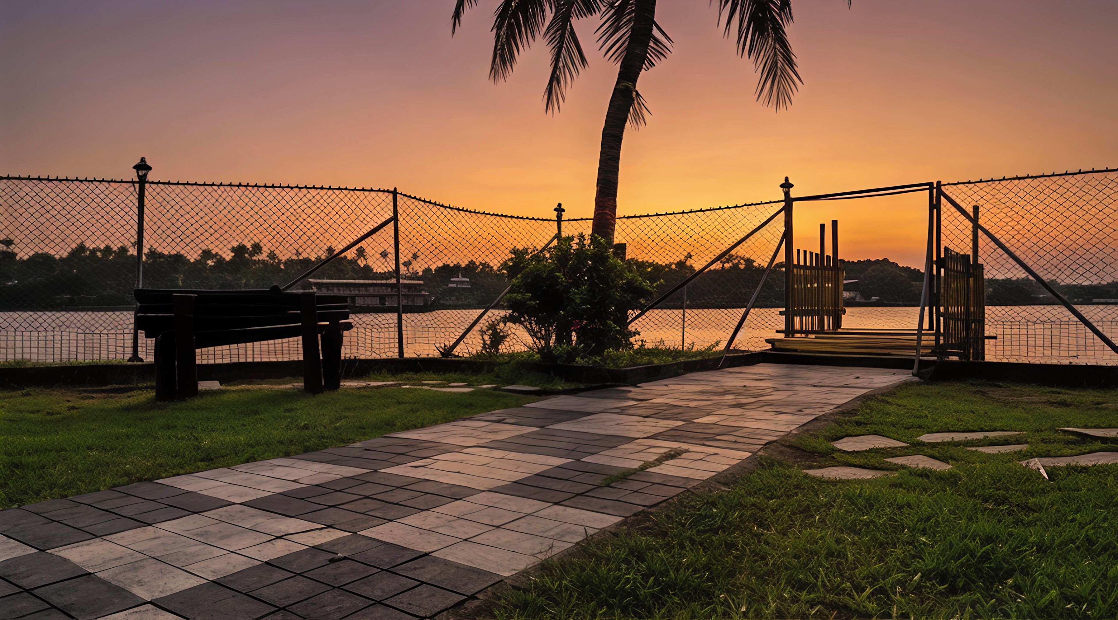 A paved walkway with a bench, a fence, and a palm tree.