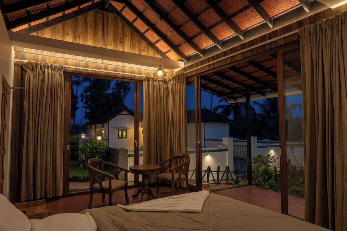 A room with a bed, two chairs, a table, and a large sliding glass door leading to an outdoor area with a view of the sky and a distant building.