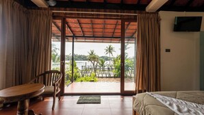 Una habitación con una cama, una mesa y una silla. Hay una puerta corrediza de vidrio que da a un área exterior con vista a palmeras y una masa de agua.