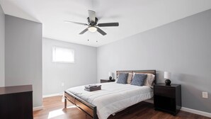 Quarto com cama, mesas de cabeceira, ventoinha no teto, janela e cômoda.