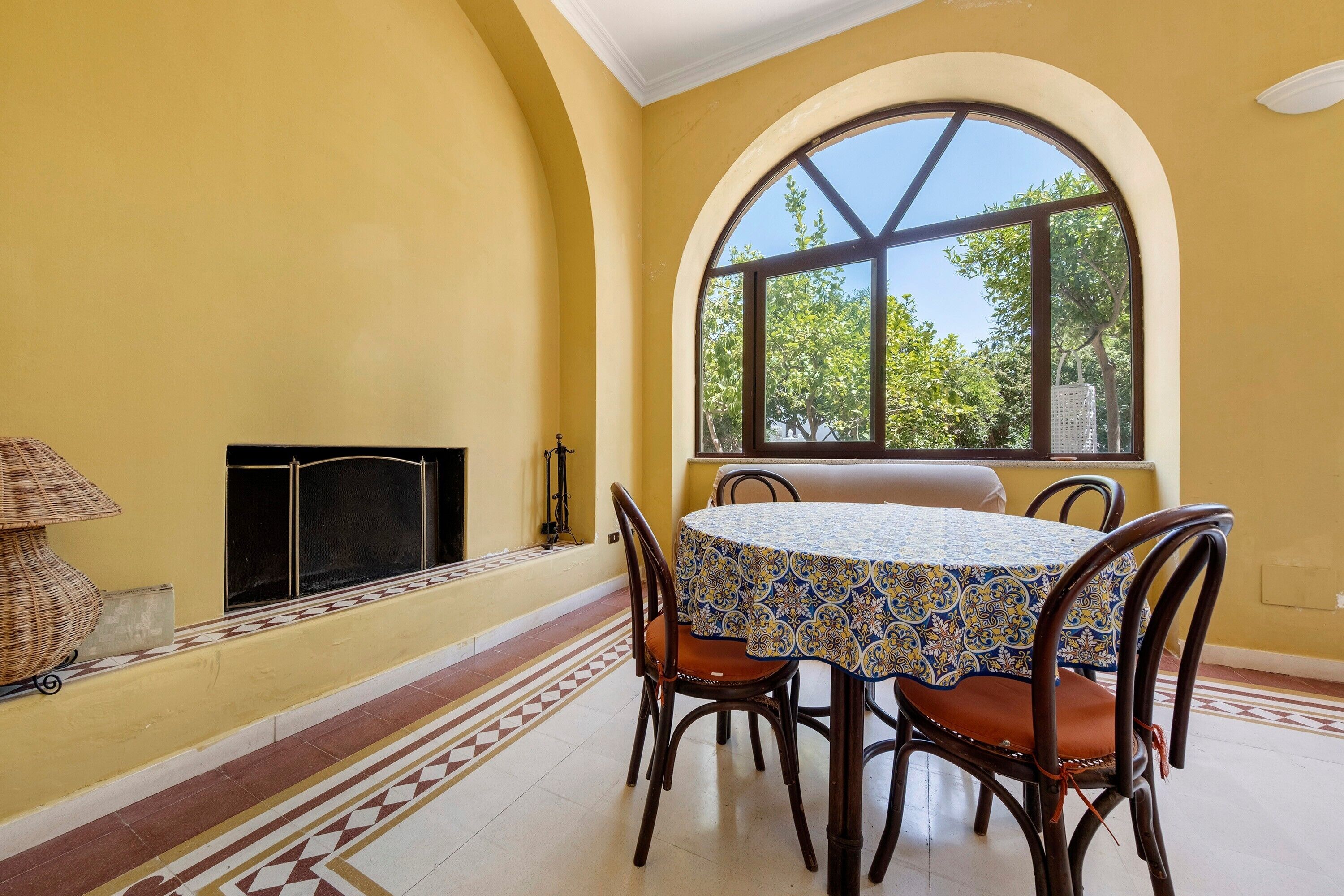 A dining area with a table and chairs, a fireplace, and a large arched window with a view of trees.