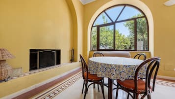 A dining area with a table and chairs, a fireplace, and a large arched window with a view of trees.