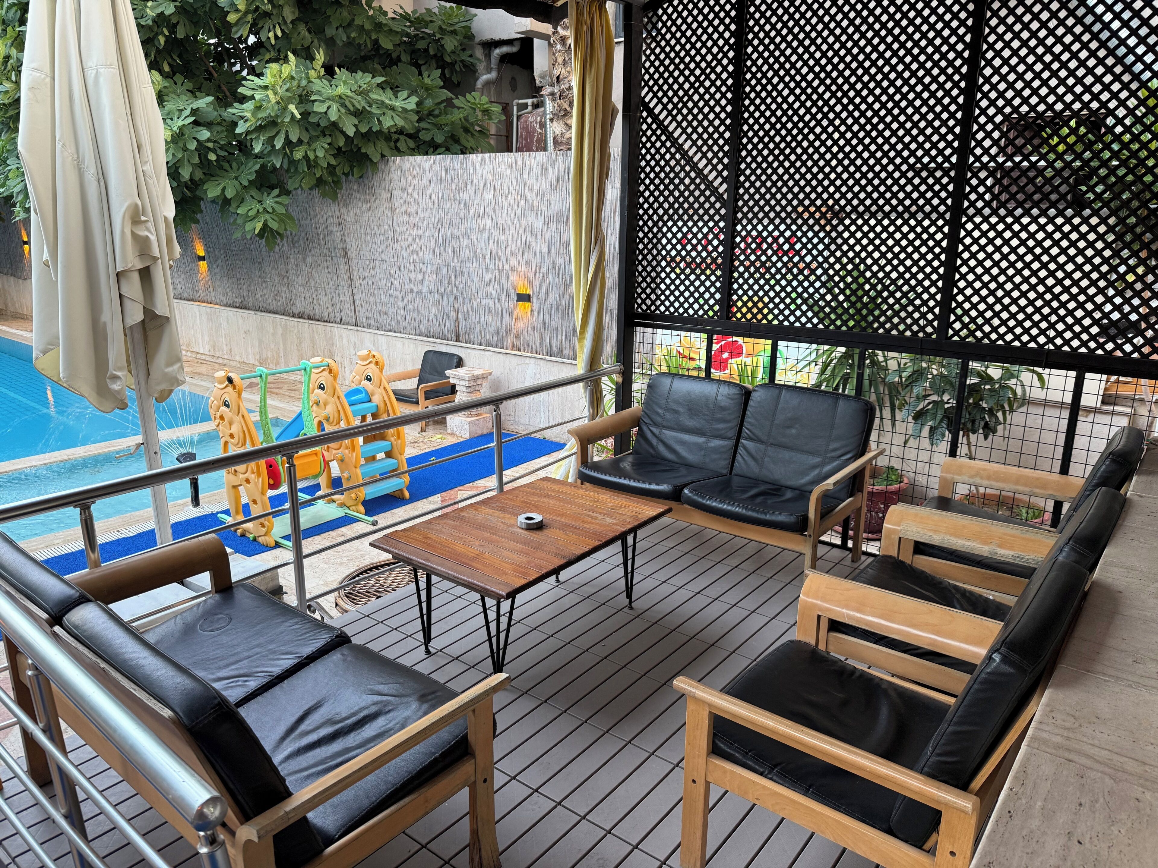 Un espacio de estar al aire libre techado con piscina, mobiliario de madera y una zona de juegos para niños.