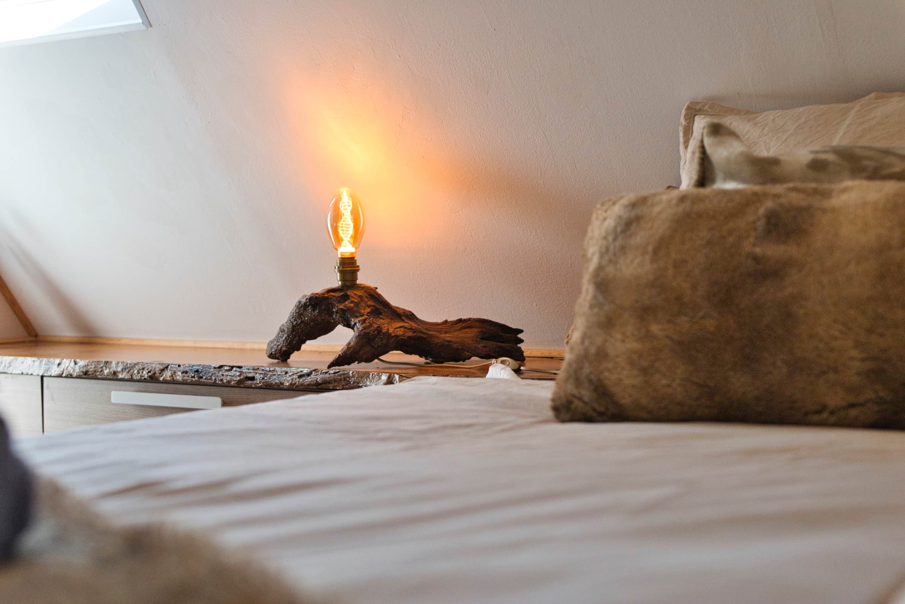 A bedroom with a bedside table, a lamp with a wooden base, and a bed with a pillow.