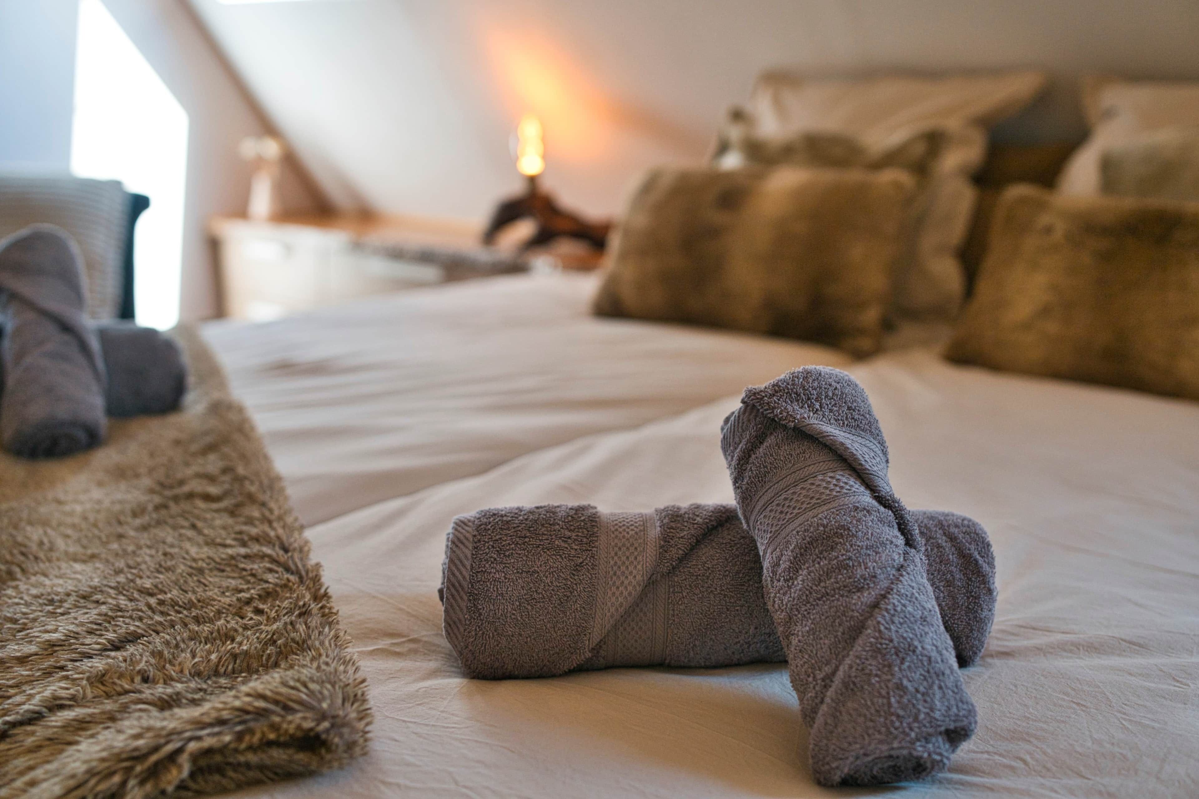 Ein ordentlich bezogenes Bett mit aufgerollten Handtüchern und einem flauschigen Teppich, was auf einen gepflegten Hotelraum hindeutet.