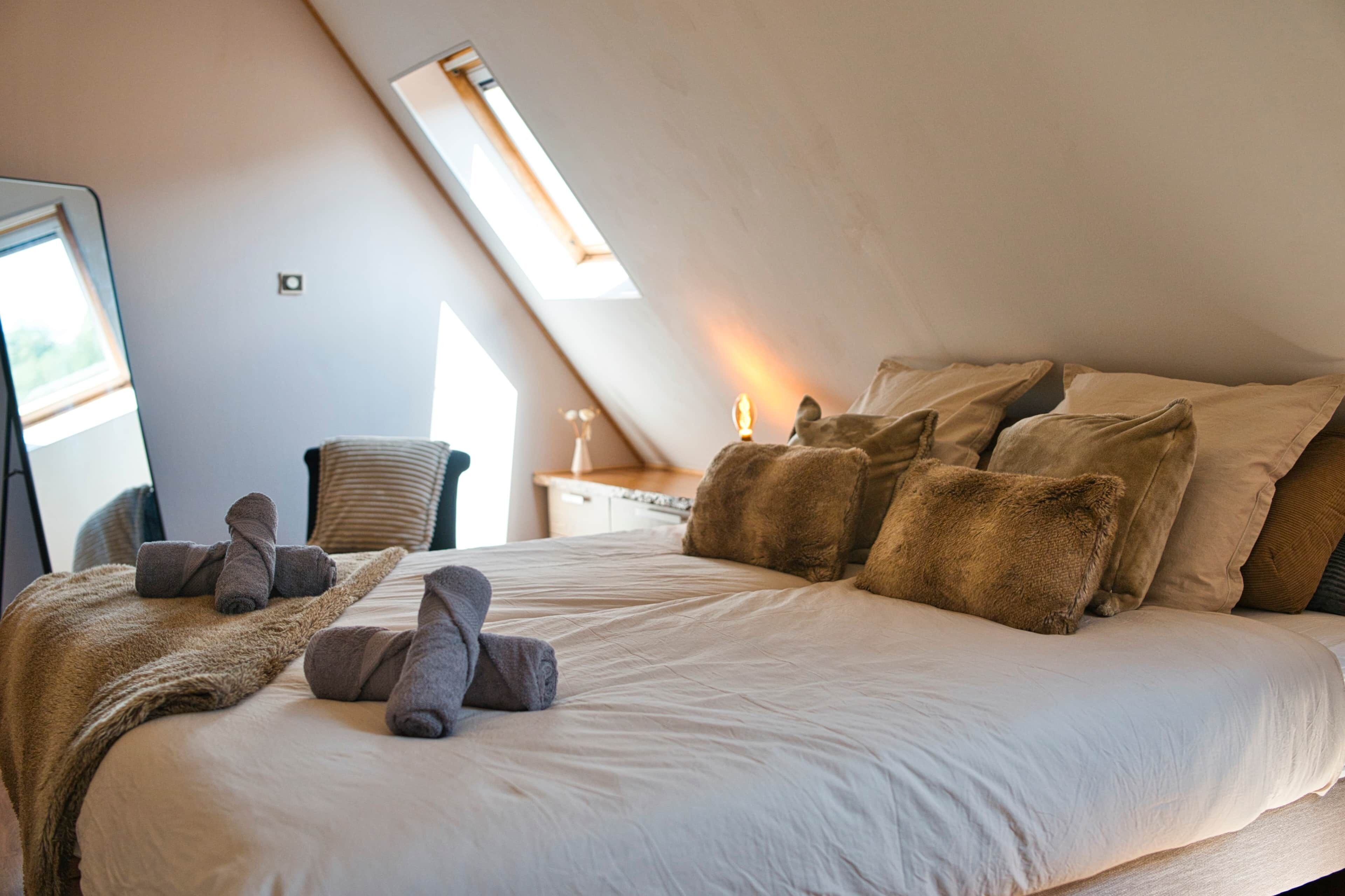 A bedroom with a large bed, multiple pillows, a chair, and a desk with a lamp.