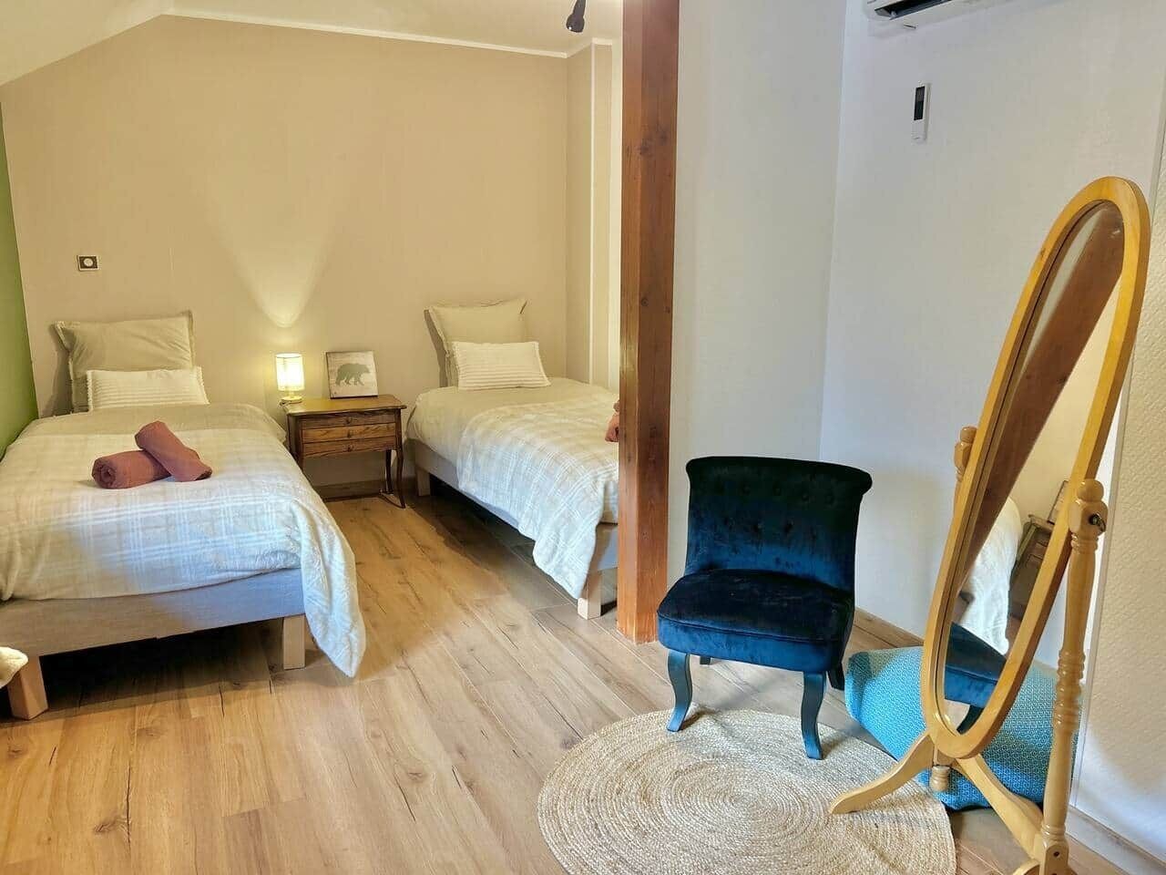 A bedroom with two beds, a wooden floor, a blue chair, and a wooden-framed mirror.