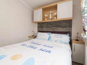 A bedroom with a bed, a nightstand, a wooden shelf with decorative items, and a headboard.