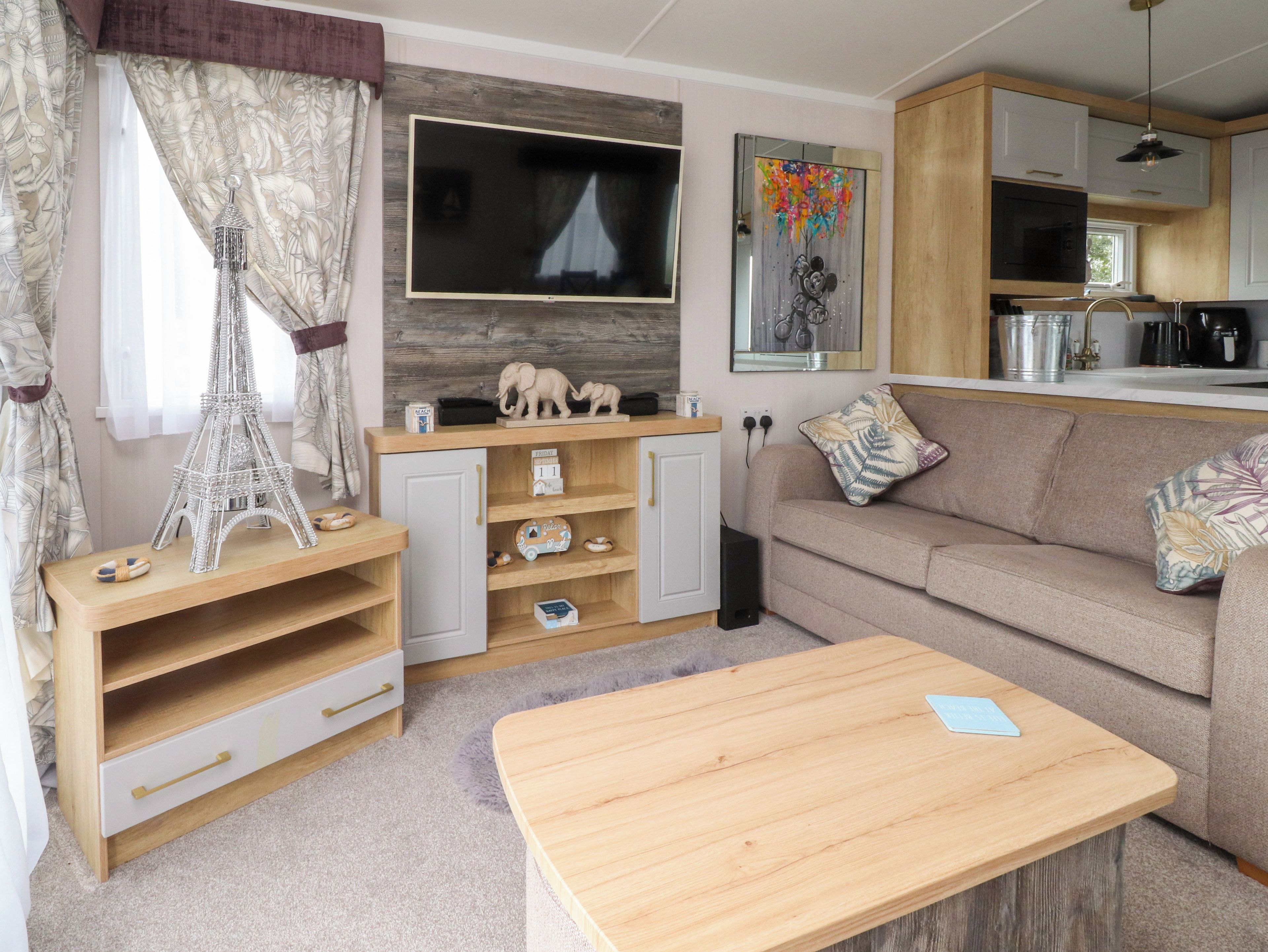 A cozy living room with a wooden coffee table, a sofa, a TV mounted on a wooden wall, and a kitchen area with cabinets and appliances.