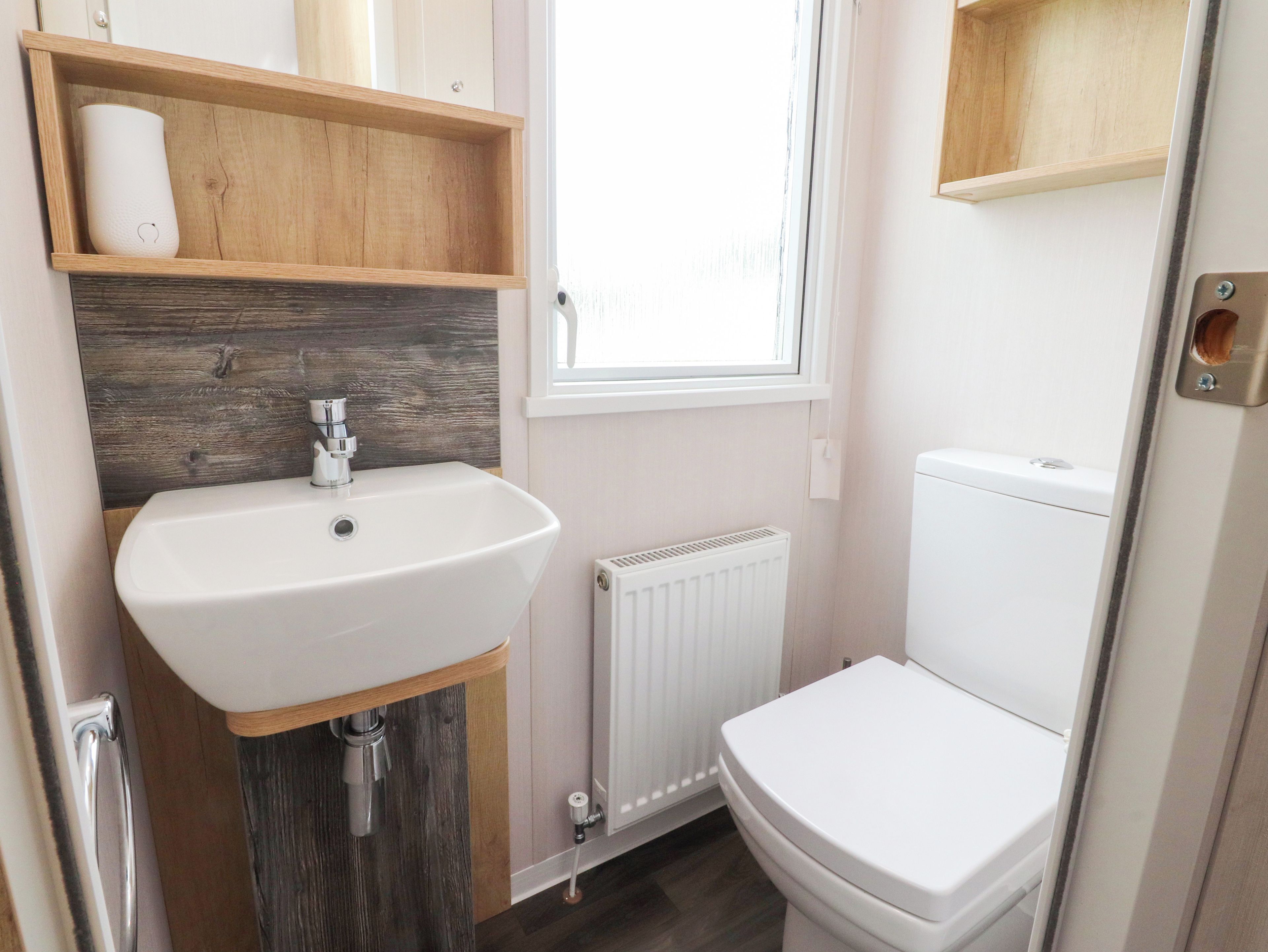 A compact bathroom with a white sink, wooden shelves, a white toilet, and a window.