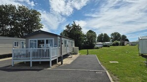 A mobile home with a porch and a small deck, surrounded by a grassy area and trees. - Marton Mere Westminster (Blackpool)