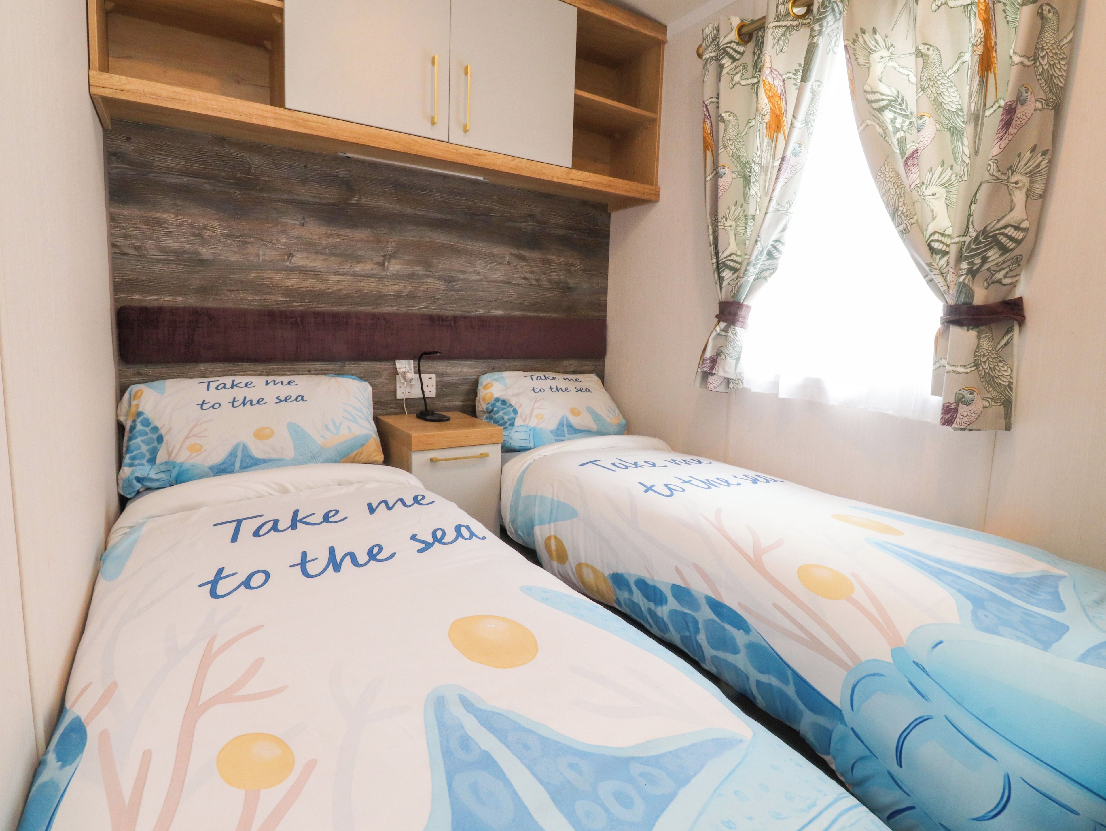 A bedroom with two beds, wooden headboard, a small wooden shelf, and a window with curtains.