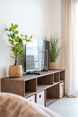 A modern living room with a flat-screen TV on a wooden console, two potted plants, and a large window with curtains. - Pilar Sunplace Deluxe, ground-floor resort apt, pool, garden view, by OYOOSTAYS (Pilar de la Horadada)