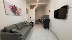 Passadís estret amb un sofà gris, una taula de dinar, televisió penjada a la paret i una cuina amb electrodomèstics.