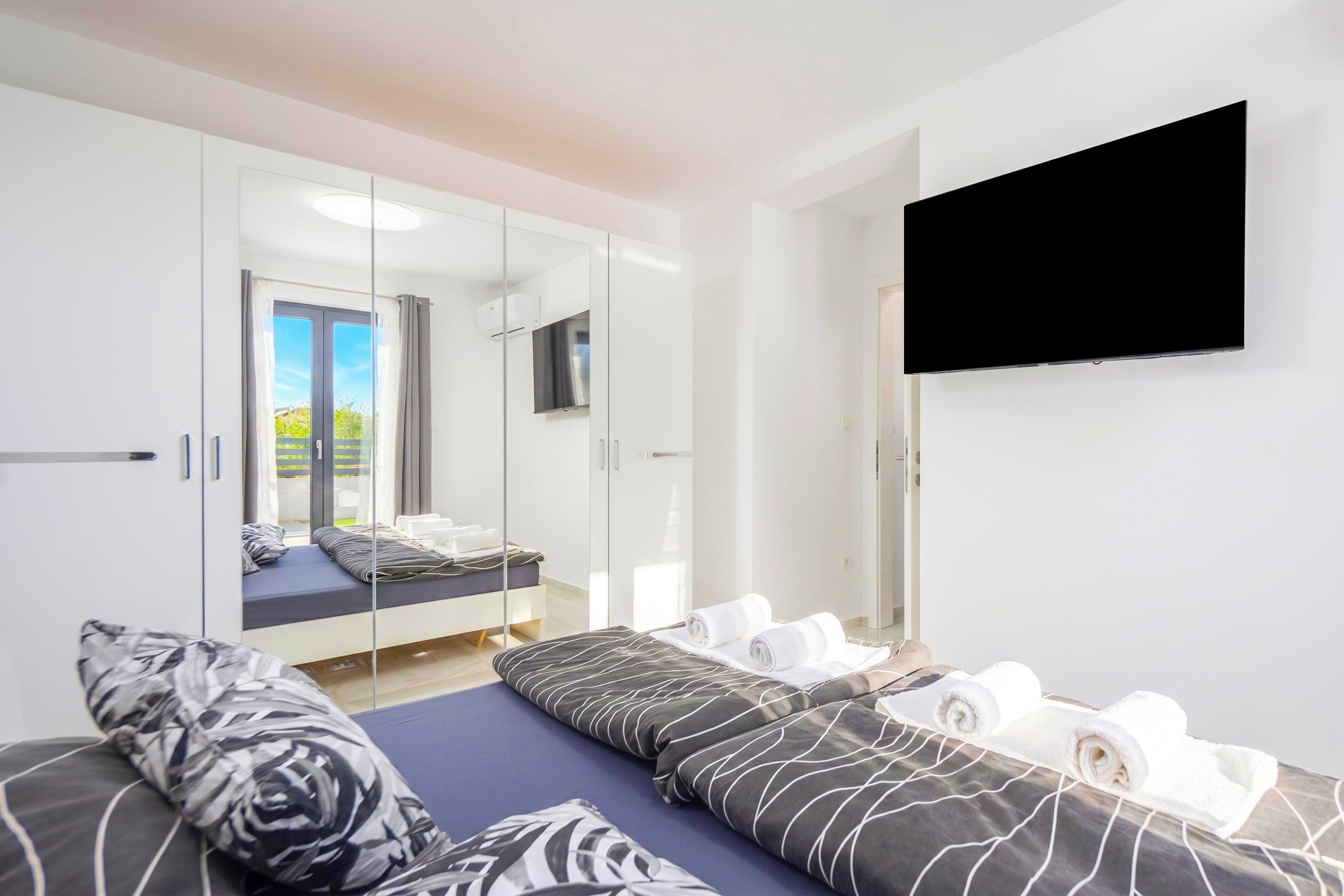 A modern hotel room with a bed, a TV, a mirror, and a view of the outdoors.
