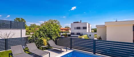 A swimming pool with lounge chairs, a fence, and modern buildings in the background.