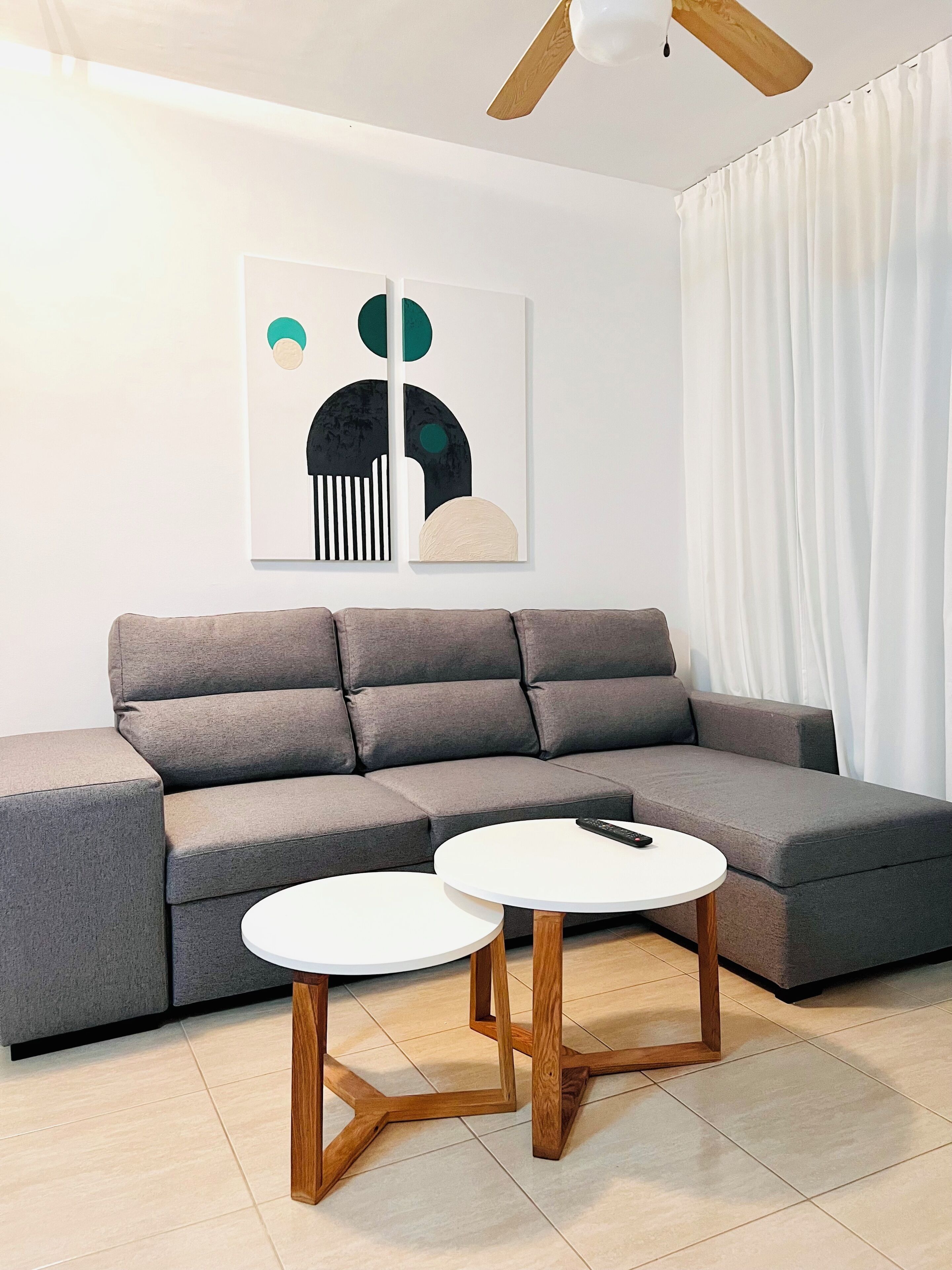Una sala de estar moderna con un sofá seccional gris, dos mesas redondas de madera y un ventilador de techo.