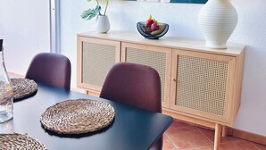 Un coin repas avec une table noire, des sets de table en fibres naturelles, un vase blanc avec une plante, un panier de fruits et une lampe posée sur un meuble en bois.