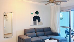 Un salon moderne avec un canapé d’angle, un ventilateur de plafond, un coin repas avec une table et des chaises, et une grande fenêtre.