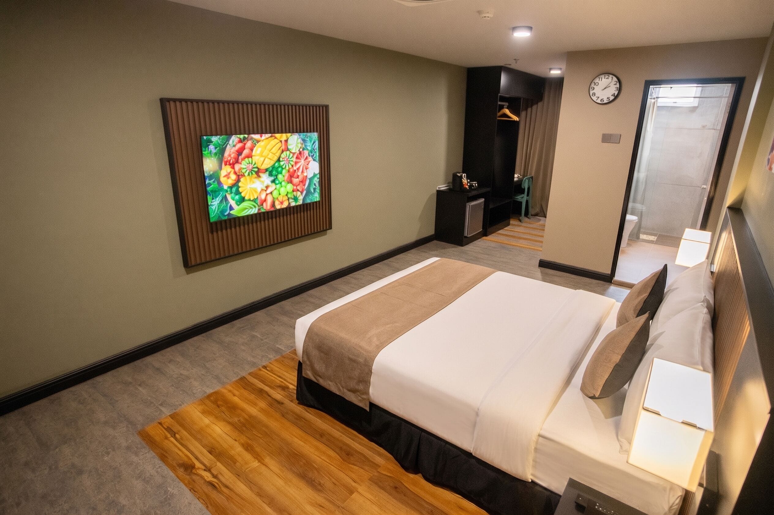Une chambre d’hôtel comprenant un lit, un téléviseur fixé au mur, un bureau avec une chaise et une horloge murale.