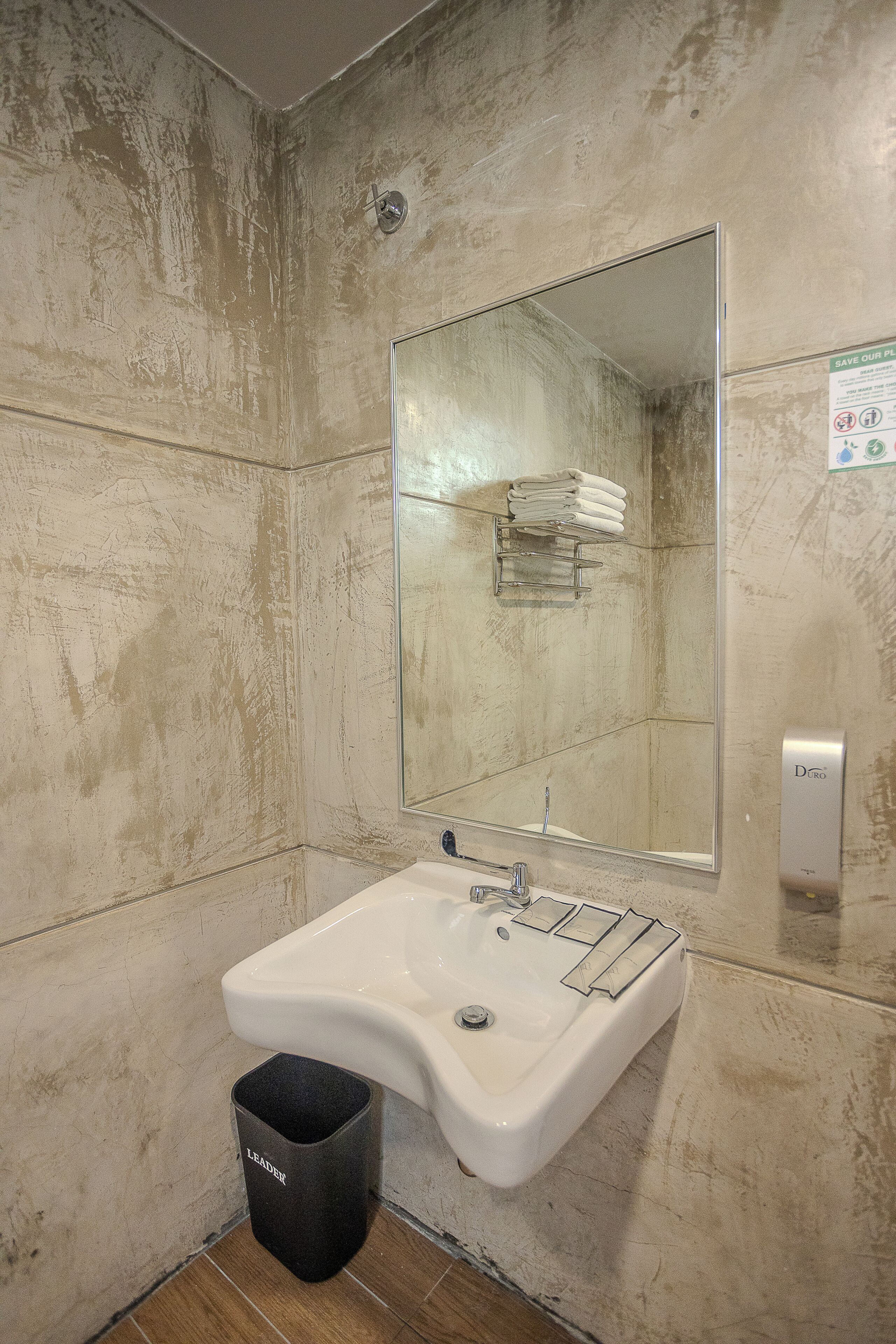 A bathroom with a white sink, a mirror, and a towel rack. There is a trash bin and a sign on the wall.
