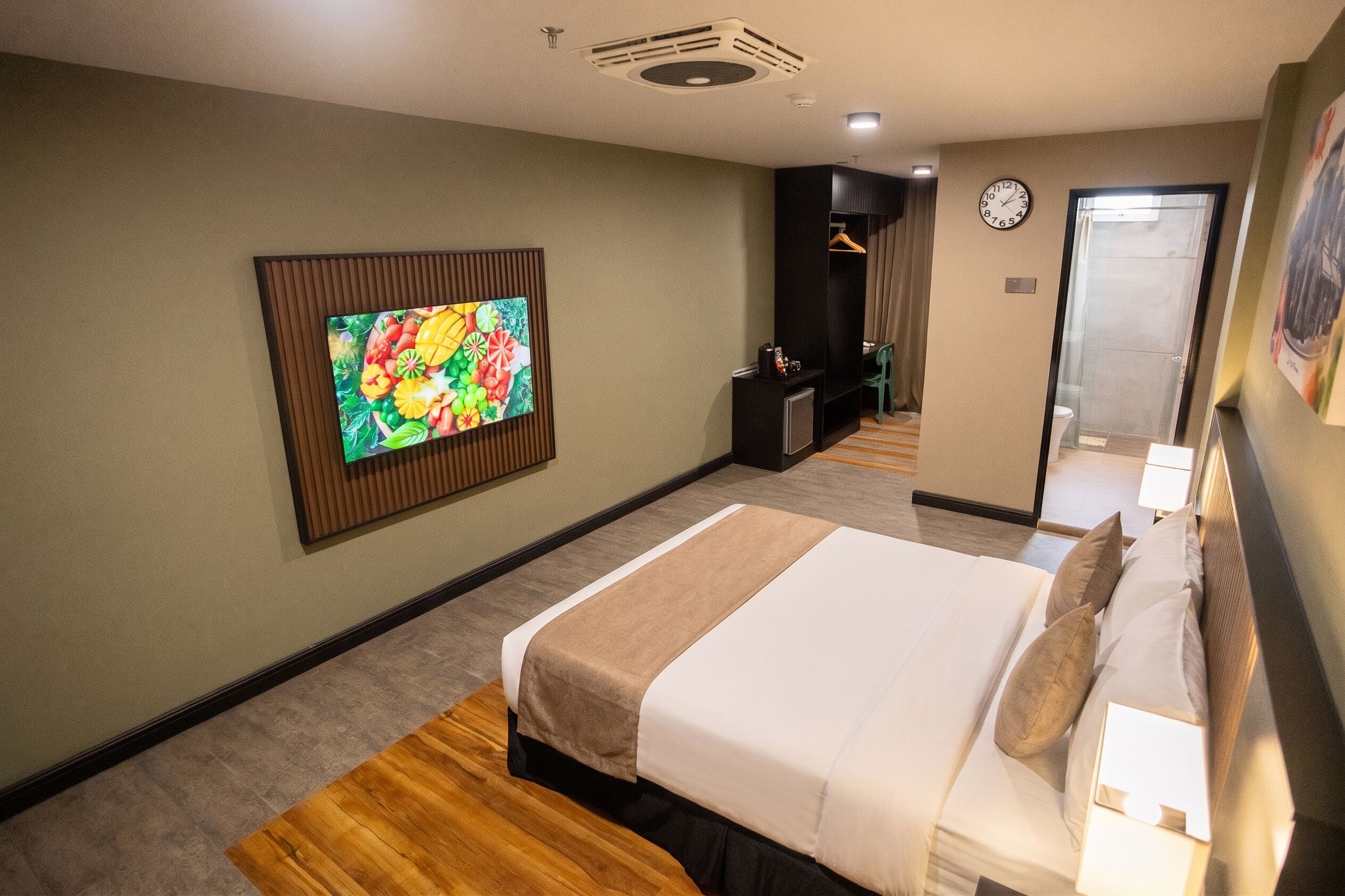 A hotel room with a bed, a television mounted on the wall, a clock, and a door leading to a bathroom.