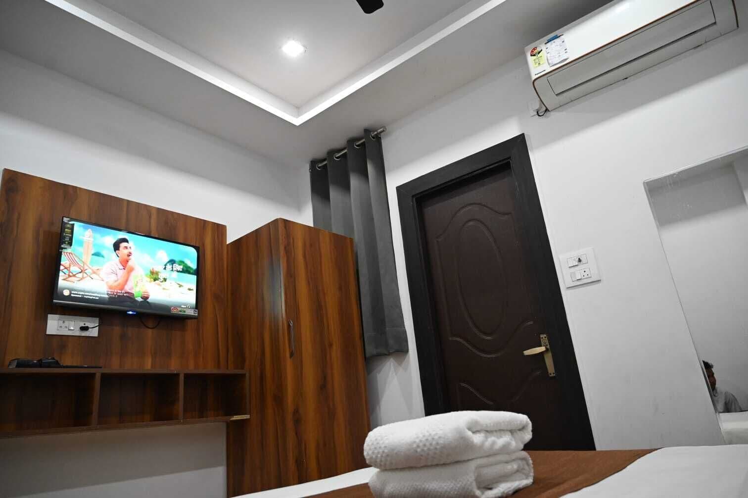 Habitación de hotel con televisión montada en la pared, armario de madera, puerta oscura y toalla blanca sobre la cama.