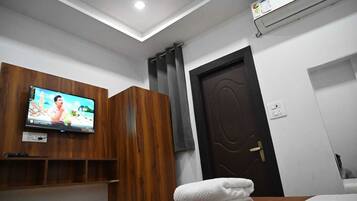 Habitación de hotel con televisión montada en la pared, armario de madera, puerta oscura y toalla blanca sobre la cama.
