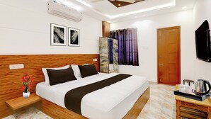 A hotel room with a bed, a ceiling fan, an air conditioning unit, a wooden headboard, a nightstand with a vase of flowers, a wall-mounted TV, and a door.