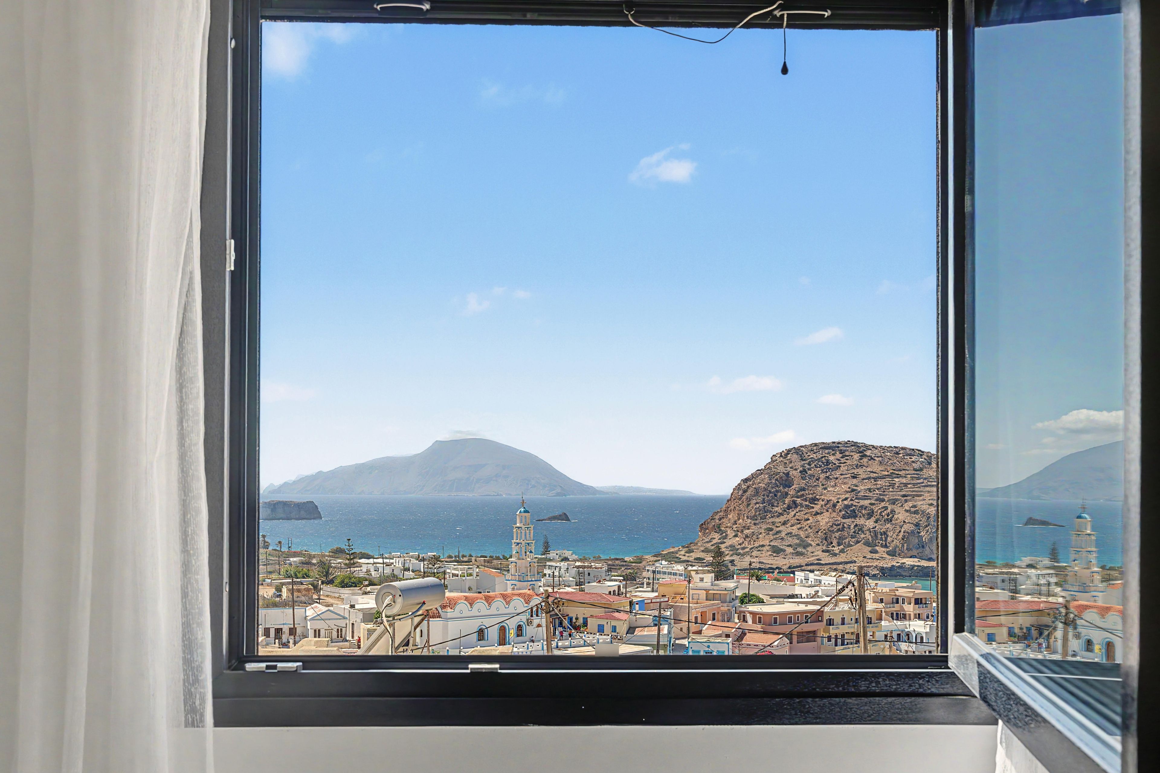 Un pueblo costero con una montaña destacada al fondo, cielo despejado y vista a través de una ventana con cortinas.