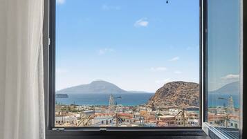 Un pueblo costero con una montaña destacada al fondo, cielo despejado y vista a través de una ventana con cortinas.