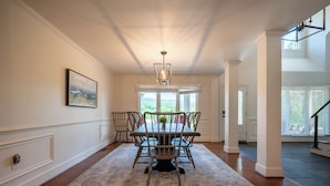 A dining area with a table and chairs, a chandelier, a view through the windows, and a staircase. - Welcome to this spectacular home, which stands out for its soaring stone fireplace, a bright sunroom, and a unique indoor-outdoor entertainment lounge with a classic arcade game! (Elliston)