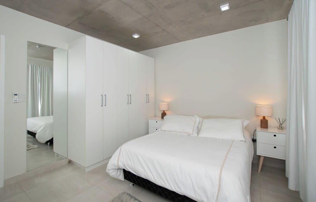 A modern bedroom with a large bed, a wardrobe, a nightstand with a lamp, and a mirror.