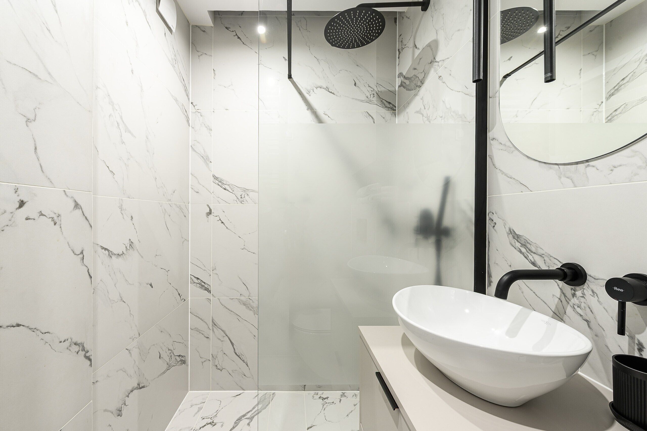 Um banheiro moderno com parede de mármore branco, box de vidro, pia escultural branca e torneira preta.