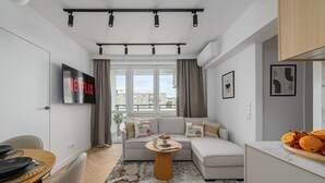 Un salon moderne avec un canapé, une table basse, un téléviseur affichant le logo « NETFLIX », et une vue sur les bâtiments à l’extérieur.
