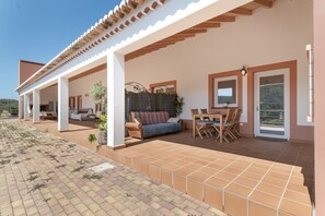 Zona exterior coberta amb un sofà, taula de dinar i cadirees, un pati enrajolat i vista al paisatge circumdant.