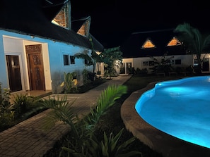 A nighttime view of a house with a swimming pool, surrounded by tropical plants and a brick pathway.
