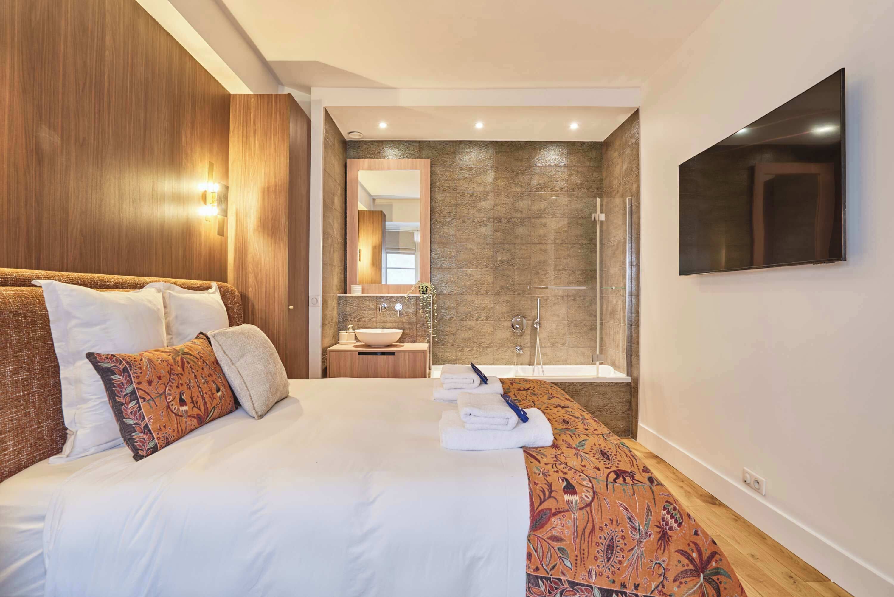 A hotel room with a large bed, a bathroom with a sink and mirror, and a flat-screen TV mounted on the wall.