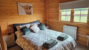 Un dormitorio de cabaña de madera con cama, almohadas, mesita de noche, lámpara, radiador y ventana con vistas al exterior.