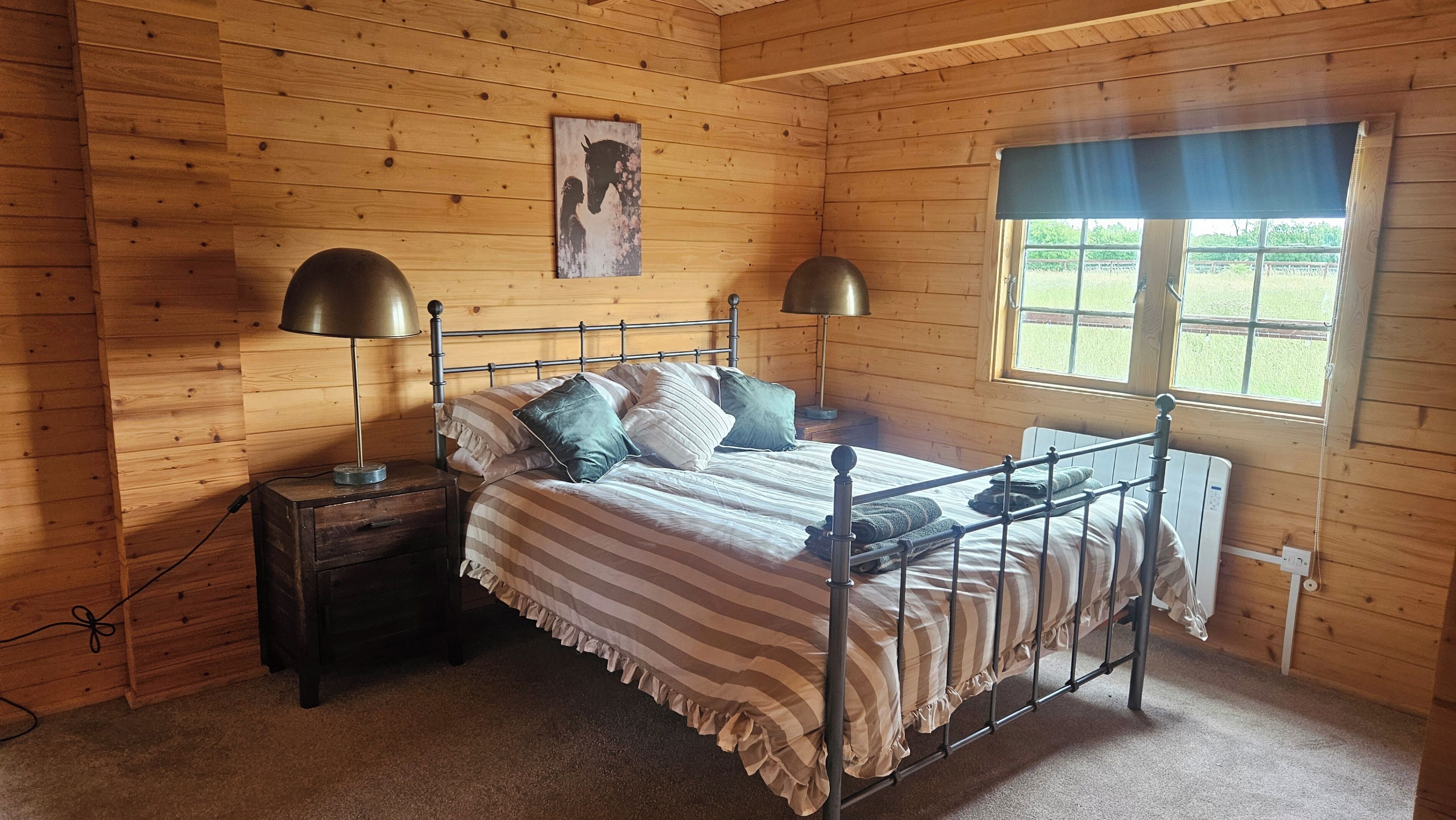 Quarto de cabana de madeira com estrutura de cama de metal, duas luminárias de cabeceira, janela com vista para vegetação e um quadro pendurado na parede.