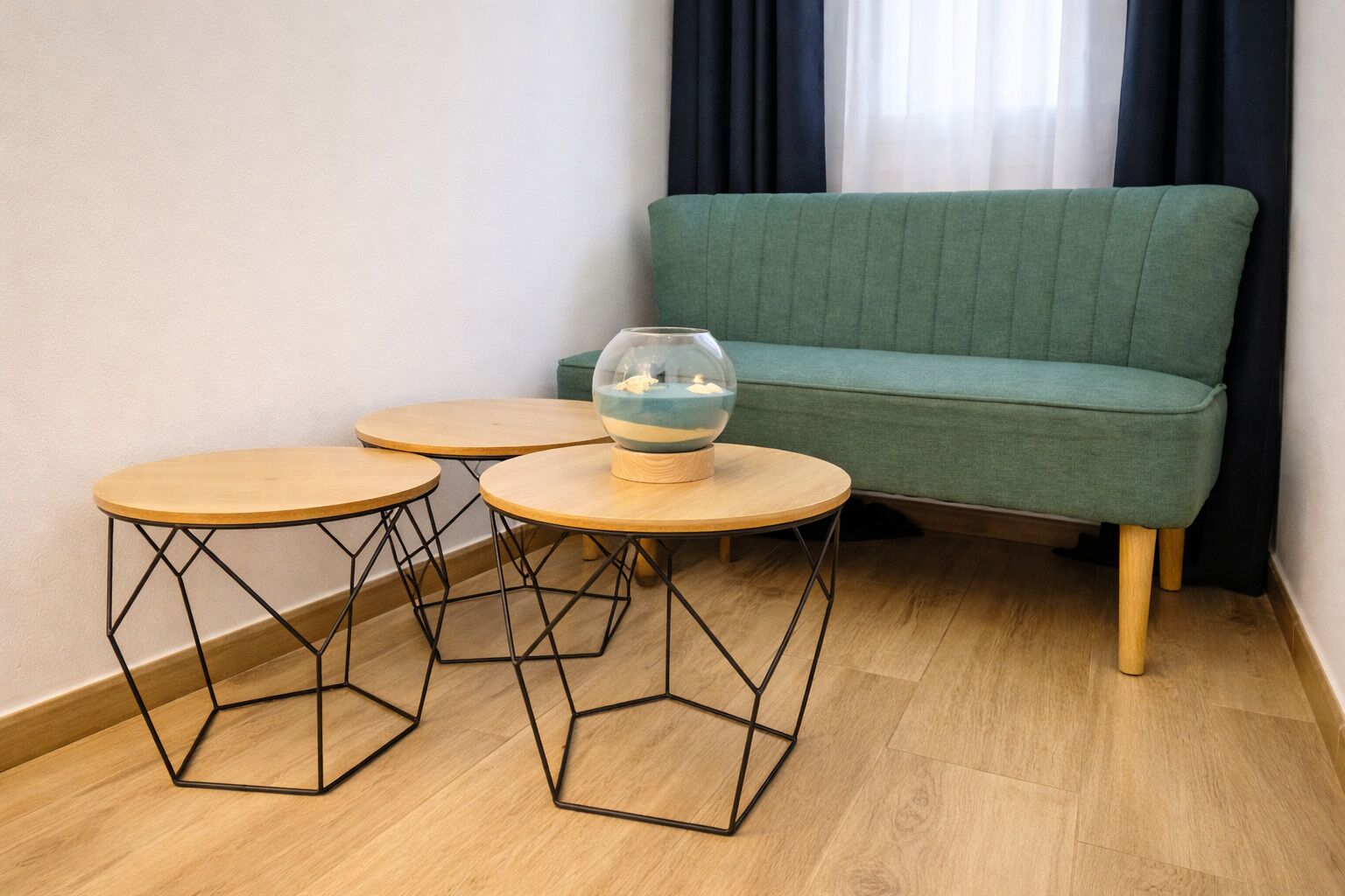 Una habitación con un sofá verde, tres mesas hexagonales con tapas de madera y estructuras de metal negro, y un cuenco de vidrio transparente sobre una de las mesas.