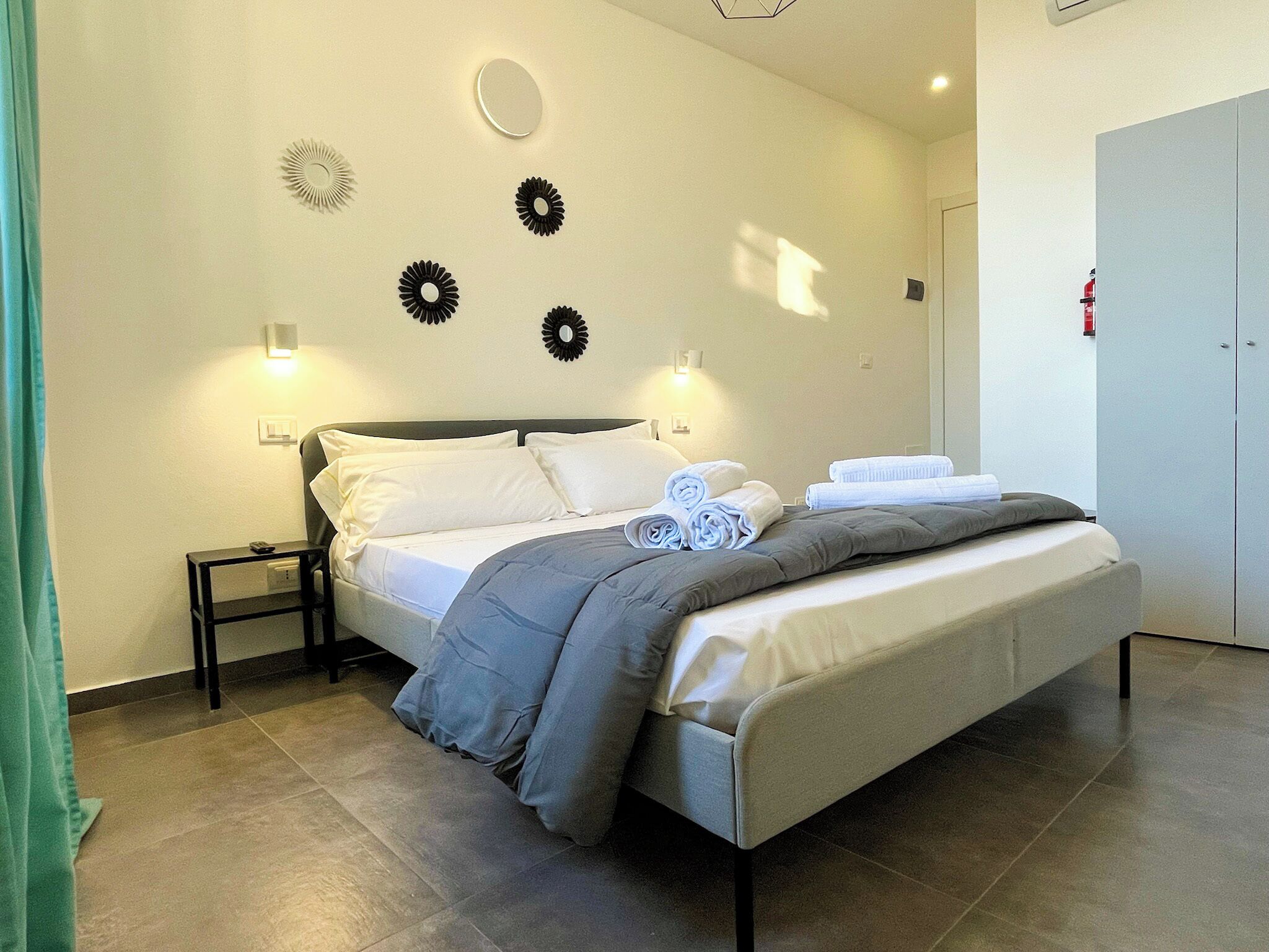 Una cama bien hecha con ropa de cama blanca y gris, una mesita de noche pequeña y apliques de luz en la pared.