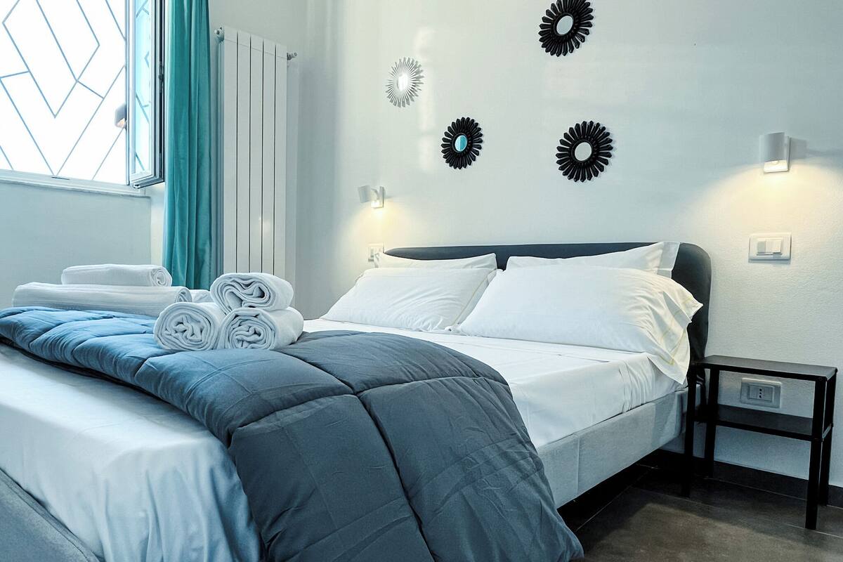 A neatly made bed with rolled towels, a dark headboard, and wall-mounted light fixtures.