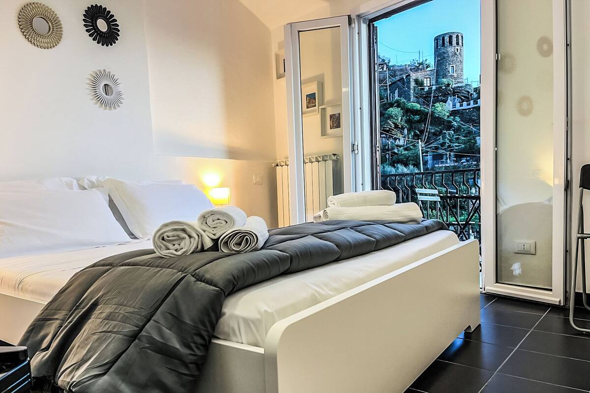A modern hotel room with a bed, a view of a cityscape, and a balcony with a railing.