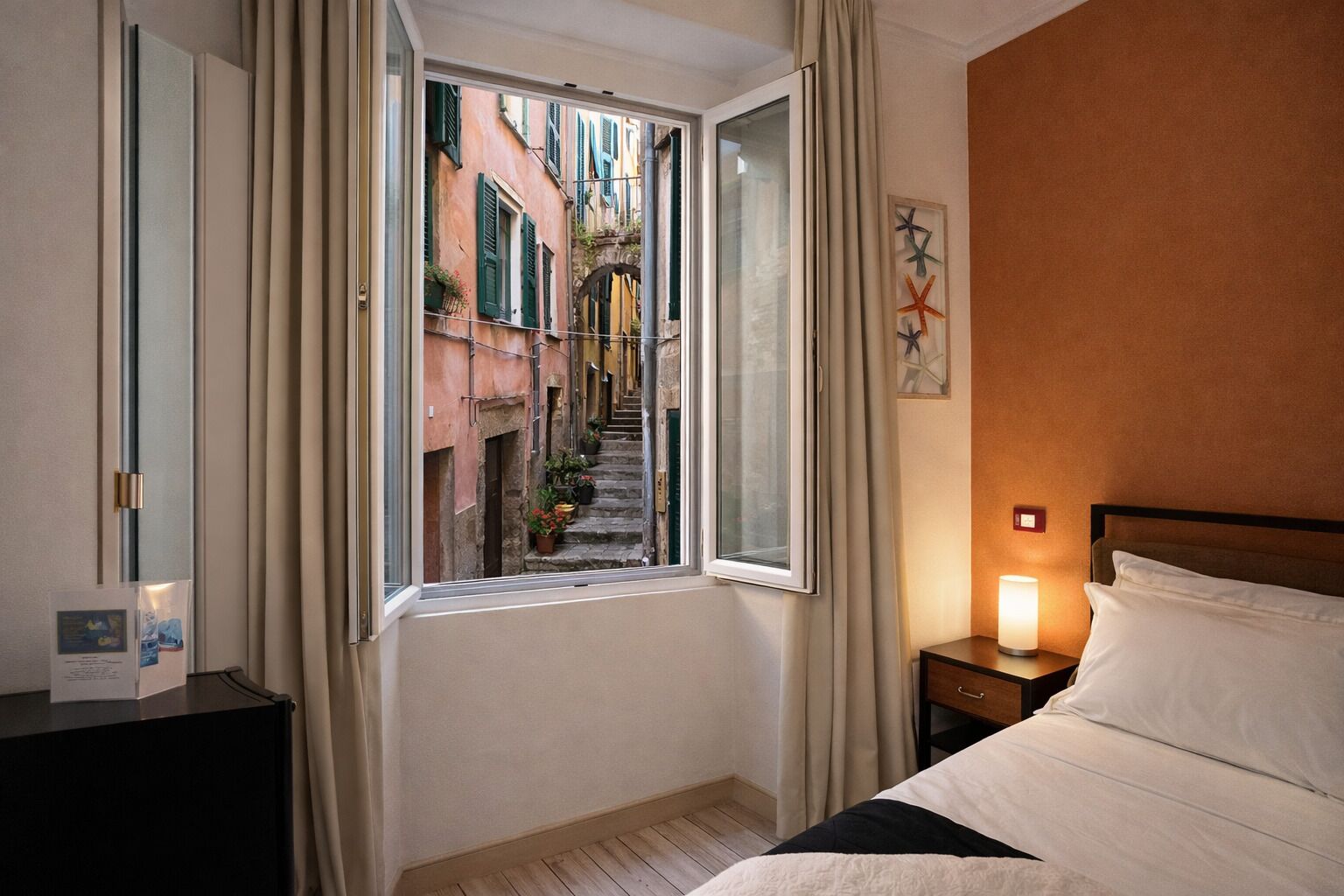Habitación de hotel con cama, mesita de noche, ventana con cortinas y vista a una calle estrecha con edificios.