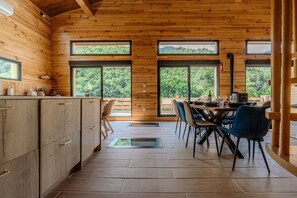 A wooden cabin interior with a dining area, kitchen, and large windows offering views of greenery.
