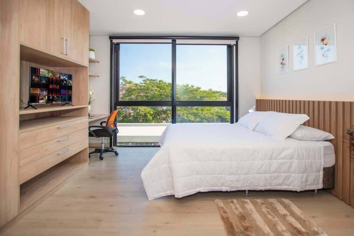 A bedroom with a bed, a desk, a TV, and a large window.